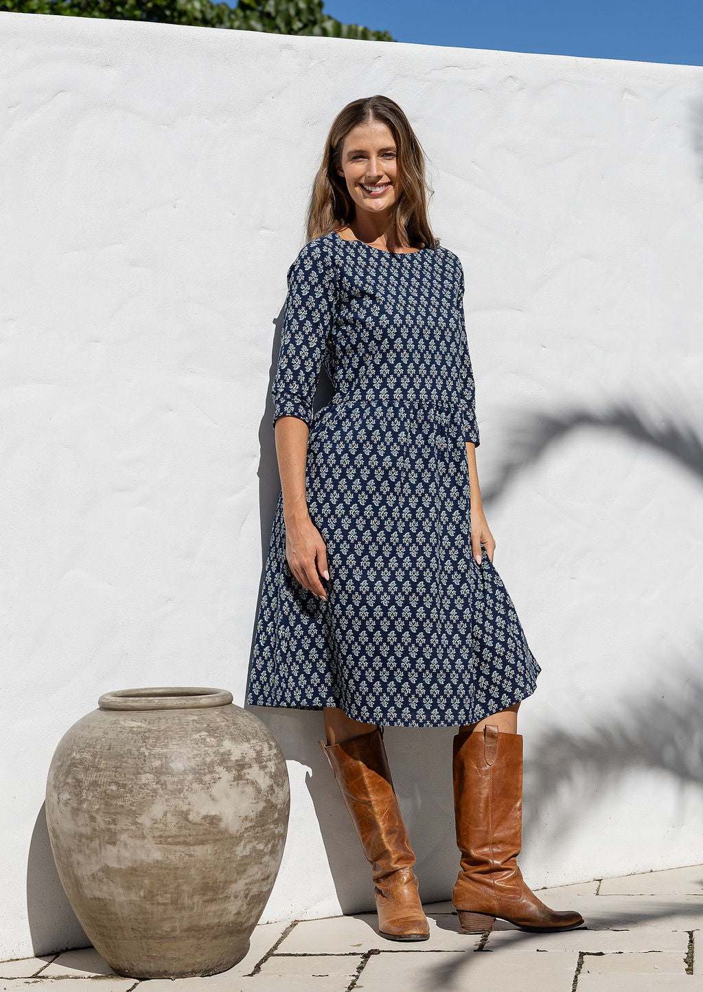 Woman in a navy blue patterned dress and brown boots standing next to a white wall