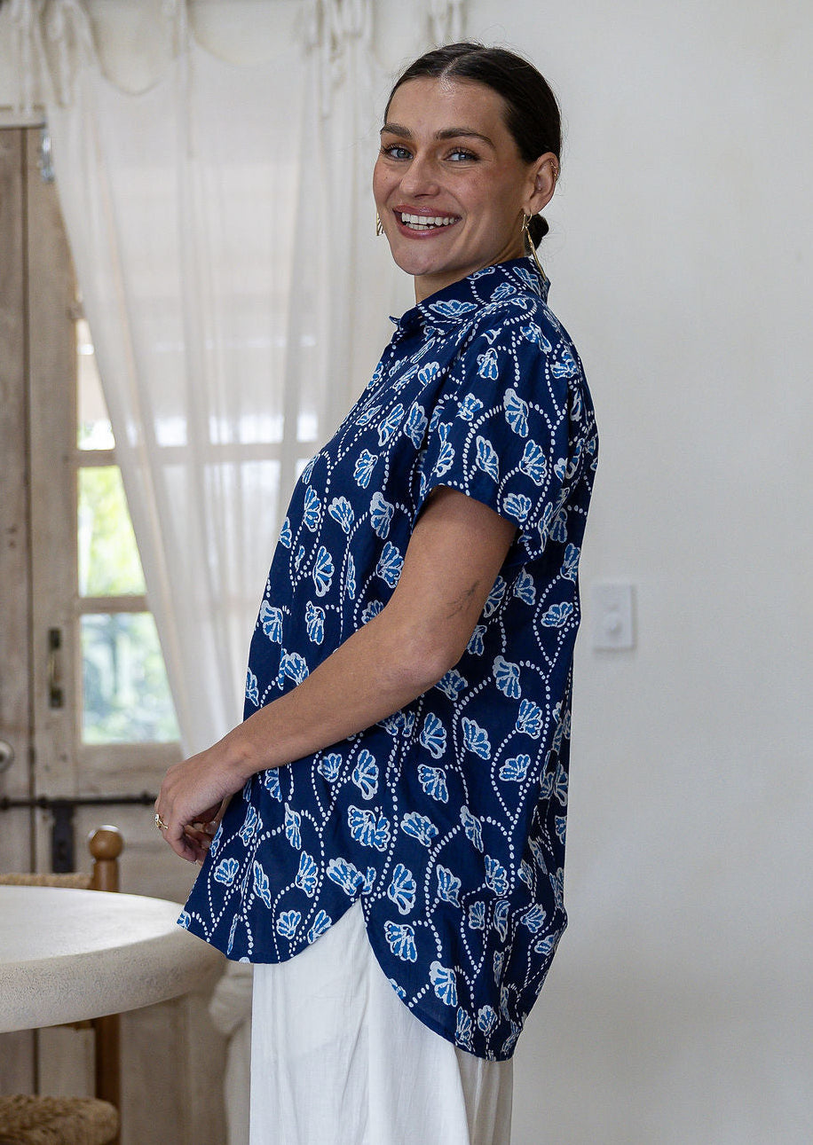 A woman standing side on wearing a navy blue cotton short-sleeve button-up shirt that is longer at the back, with a white leaf block print pattern, paired with white pants