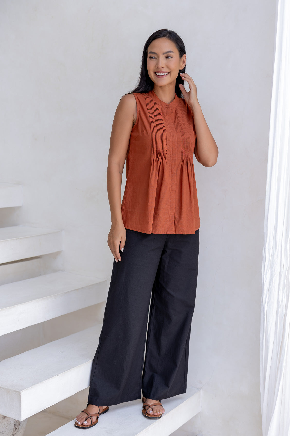 Woman wearing a rust orange sleeveless cotton top with decorative pleats and high round neckline, paired with black cotton pants.