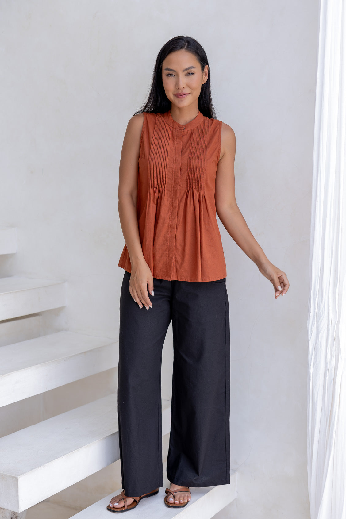 Woman wearing a rust-colored sleeveless cotton top and dark pants standing on a white staircase.