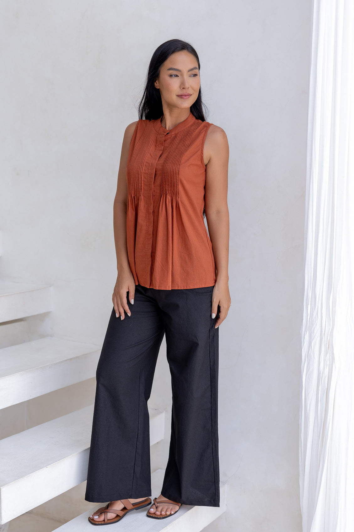 Woman wearing a fitted sleeveless cotton top in rust shade with small pin tuck detailing at the front and back.