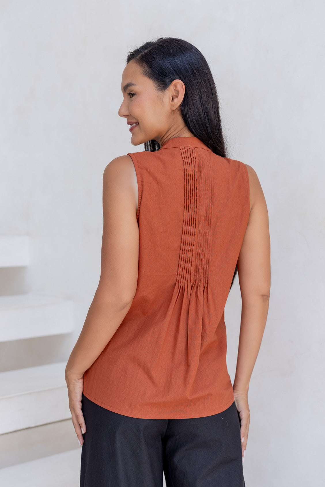 Woman wearing a 100% cotton rust-colored sleeveless top with pleated details and mandarin collar.