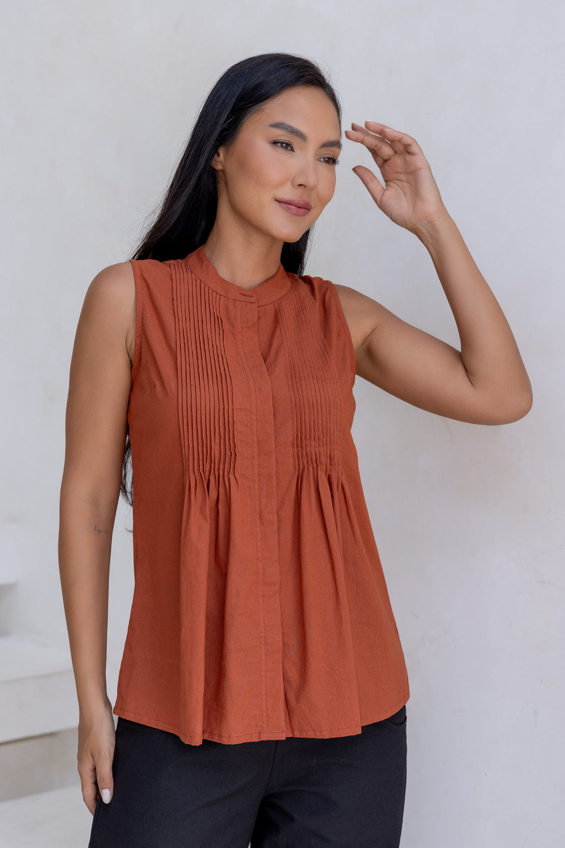 Woman wearing a 100% cotton rust coloured sleeveless top with small pleated design, and buttoned mandarin collar.