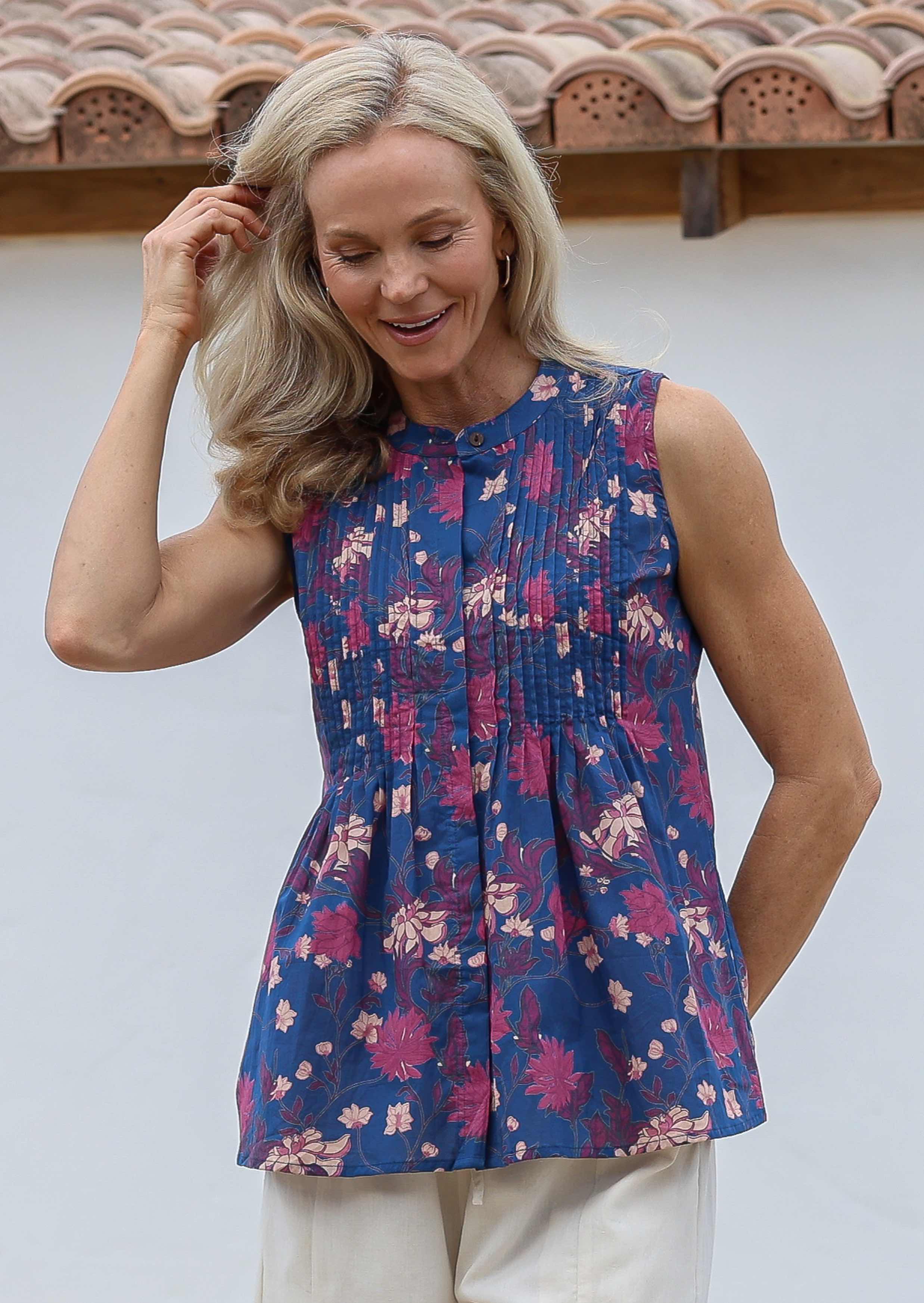 Woman posing in front of a white wall wearing a cotton sleeveless top in blue based floral print with small decorative pleats in the front.