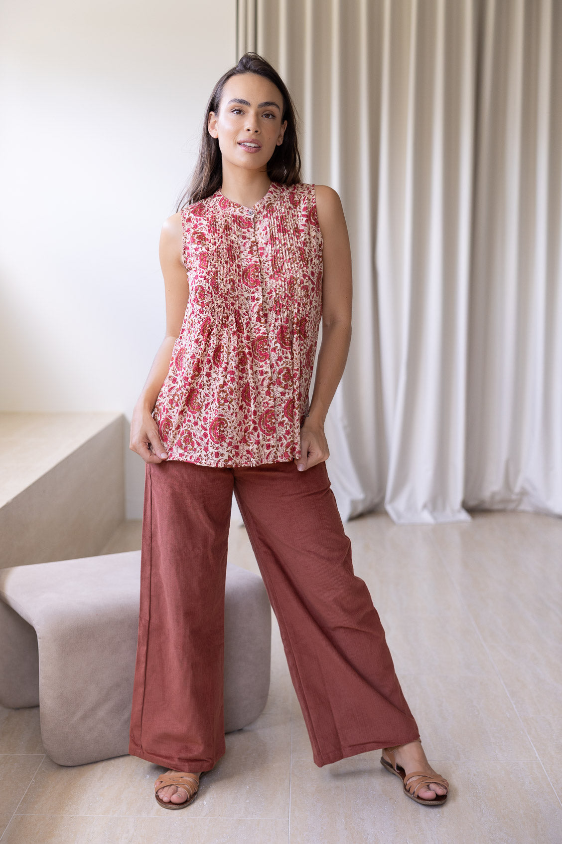 Woman wearing a floral sleeveless top with decorative pleats, high round neckline and plain pants in a room setting.