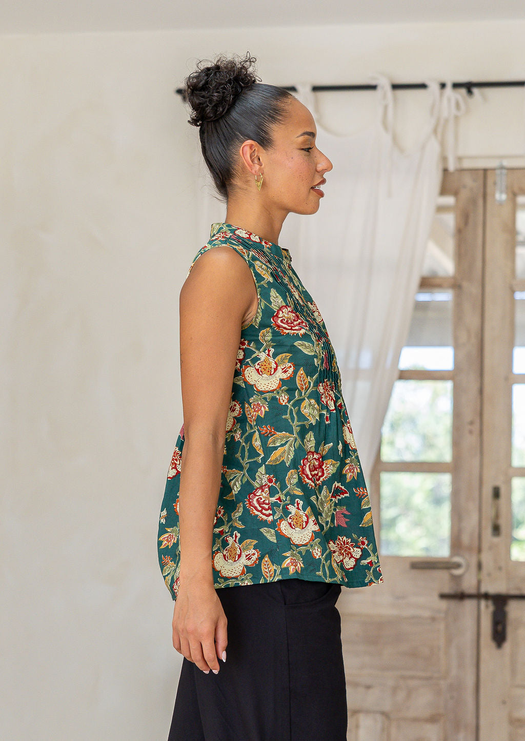 Woman stands sideways wearing a green based floral print sleeveless cotton top with buttoned mandarin collar, high round neckline and flared fit