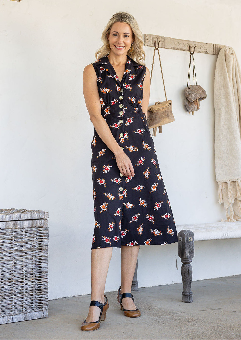 Woman wearing midi length, sleeveless cotton dress with collar and pockets in black based floral print.
