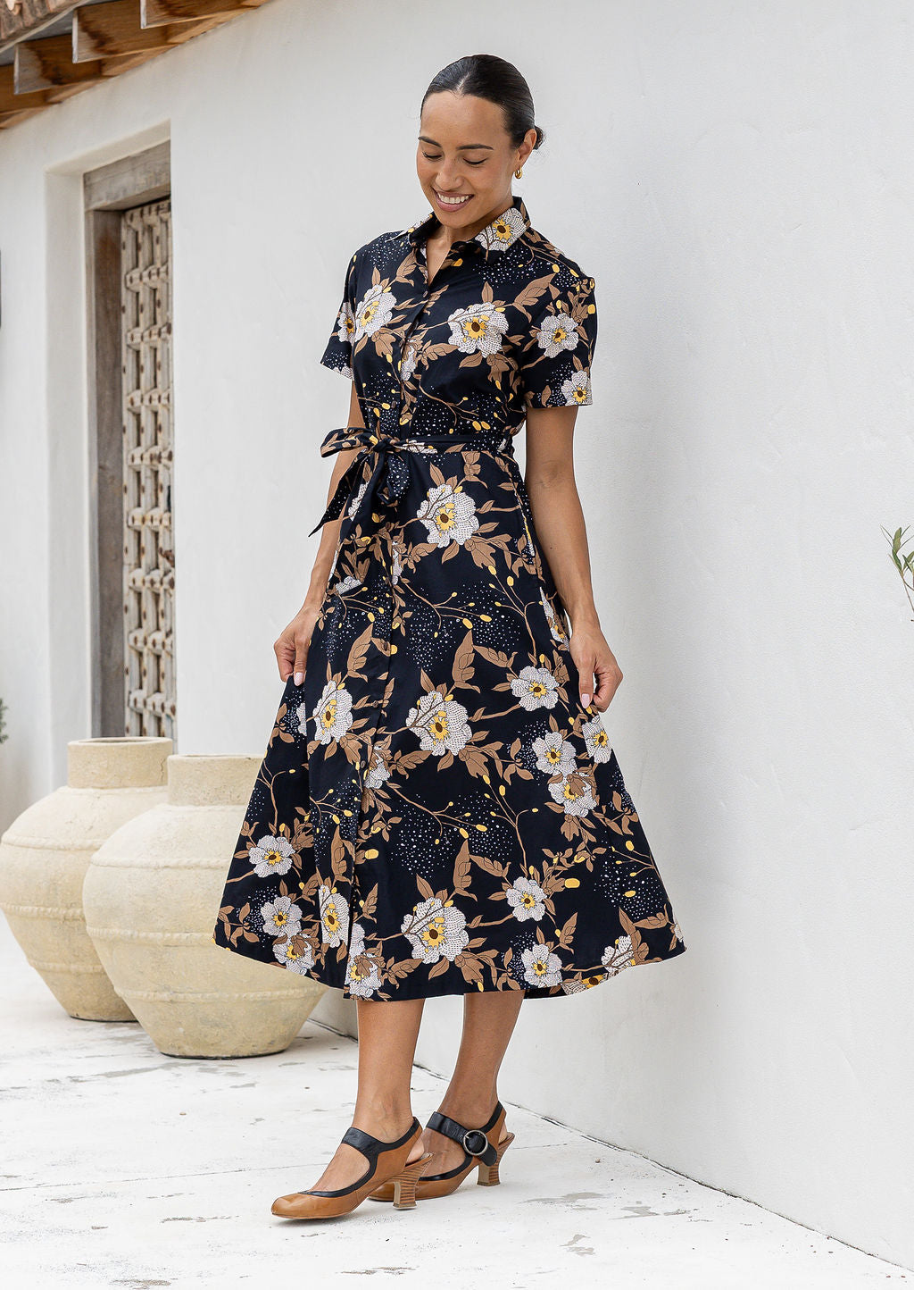 Model standing in front of white wall in floral printed shirt dress with detachable fabric belt, short sleeves and pockets.