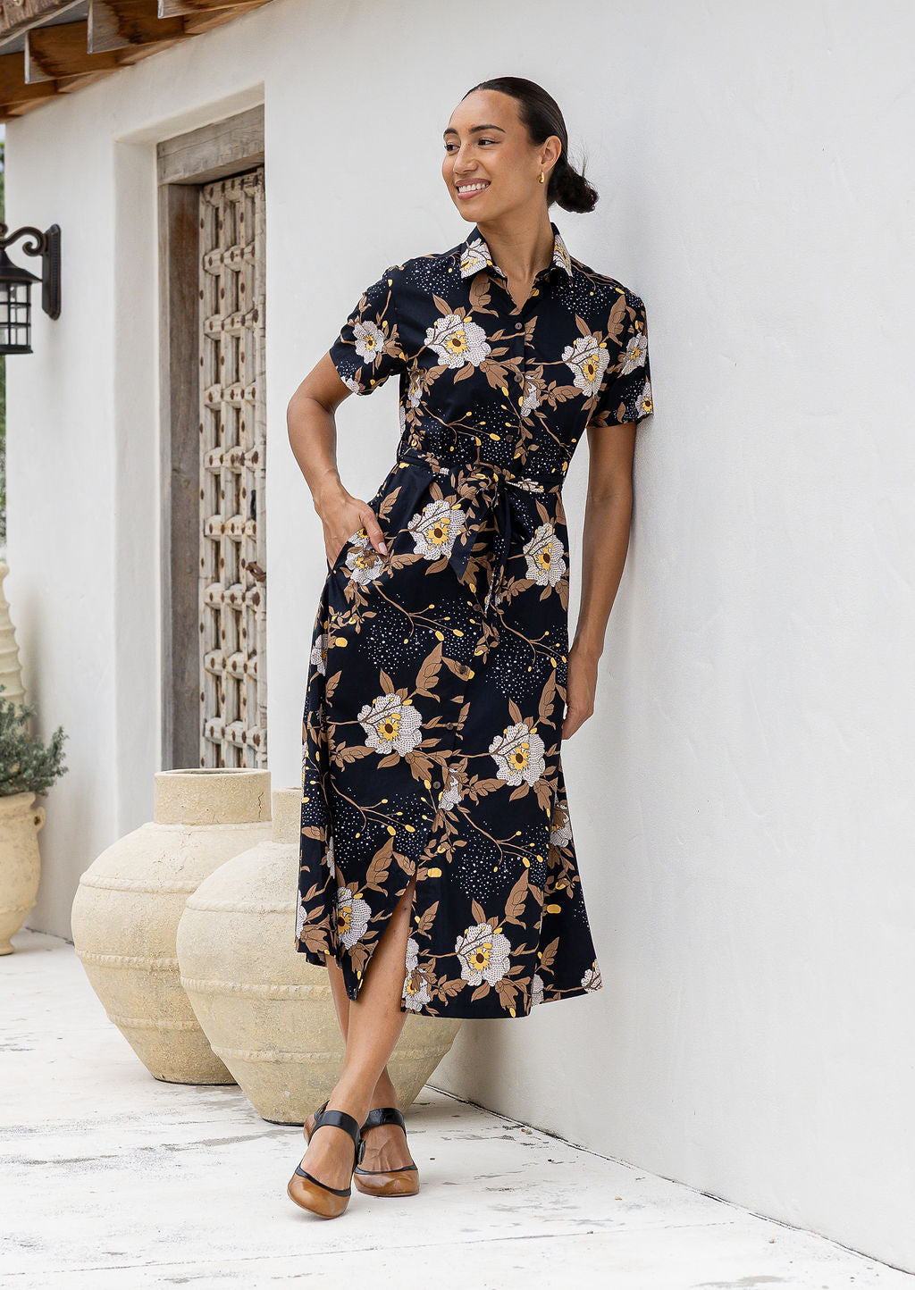 Model poses with one hand in pocket wearing a shirt style button through cotton dress with collar and a removable waist tie.