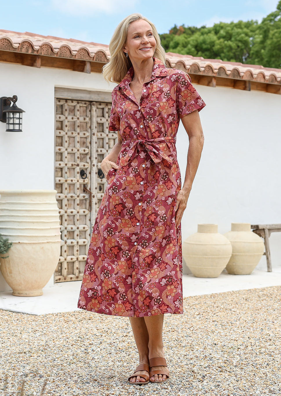 Woman wears rust pink based floral cotton shirt-style dress with collar, fabric waist tie and hidden side pockets.