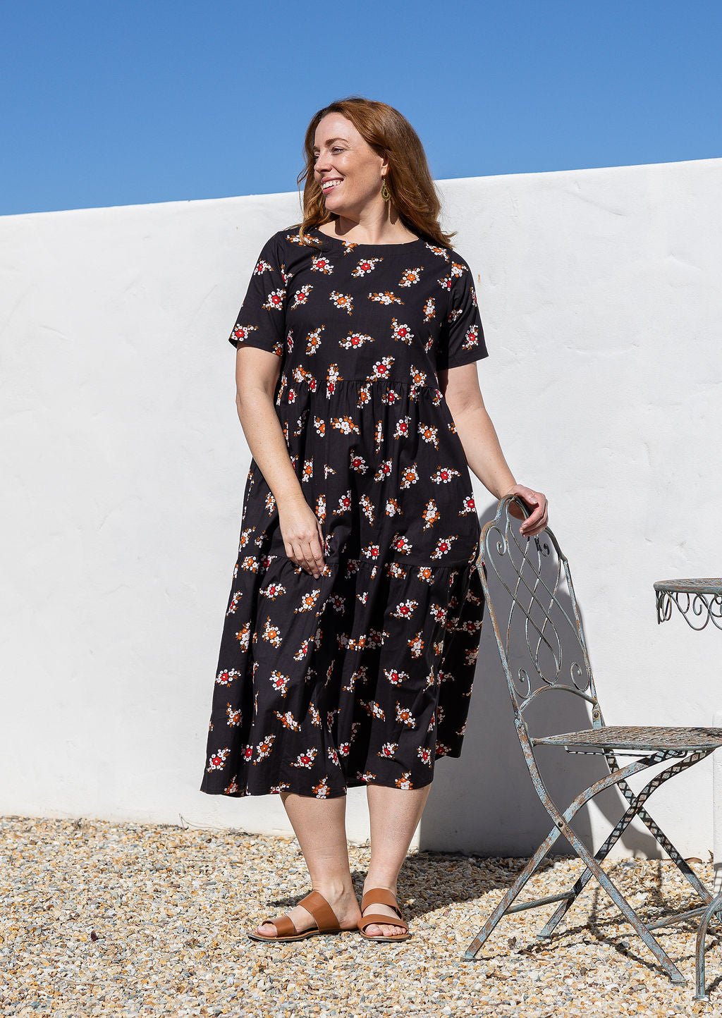 Woman standing wears a black based floral print cotton dress with three tiered design, short sleeves and hidden side pockets