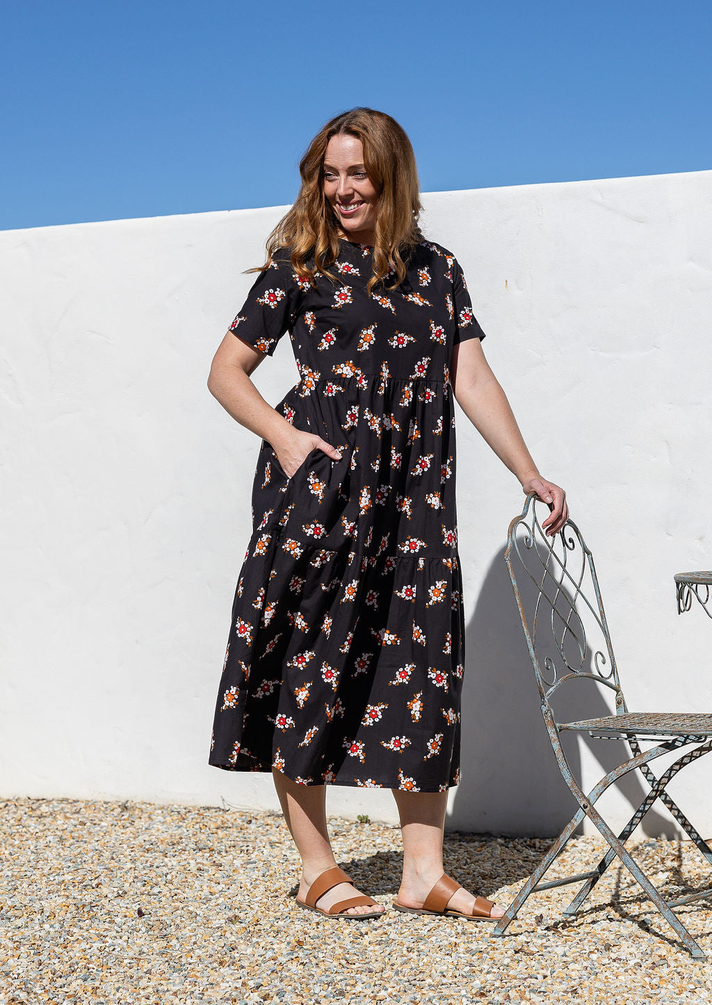Woman is wearing a relaxed fit midi length, tiered cotton dress in black based floral print with short sleeves, round neckline and side pockets.