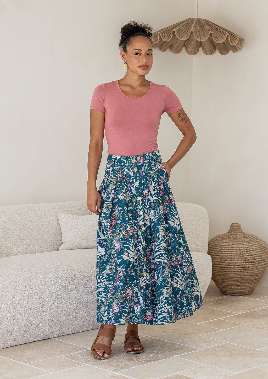 Woman wearing a pink top and green floral cotton floral maxi skirt with pockets in a room with a white couch and woven basket.