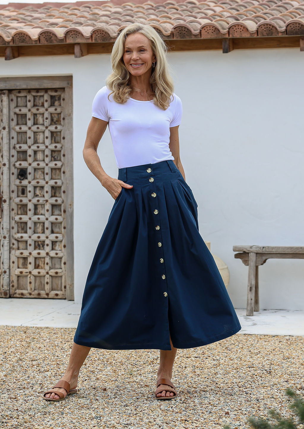 Woman wearing a white top and navy cotton skirt with buttons, belt loops and box pleats, standing in front of a white wall and wooden door