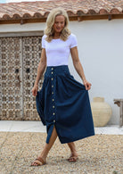 Woman wearing a white top and navy cotton, shin-length skirt with button through front, belt loops and pockets, standing in front of a wooden door