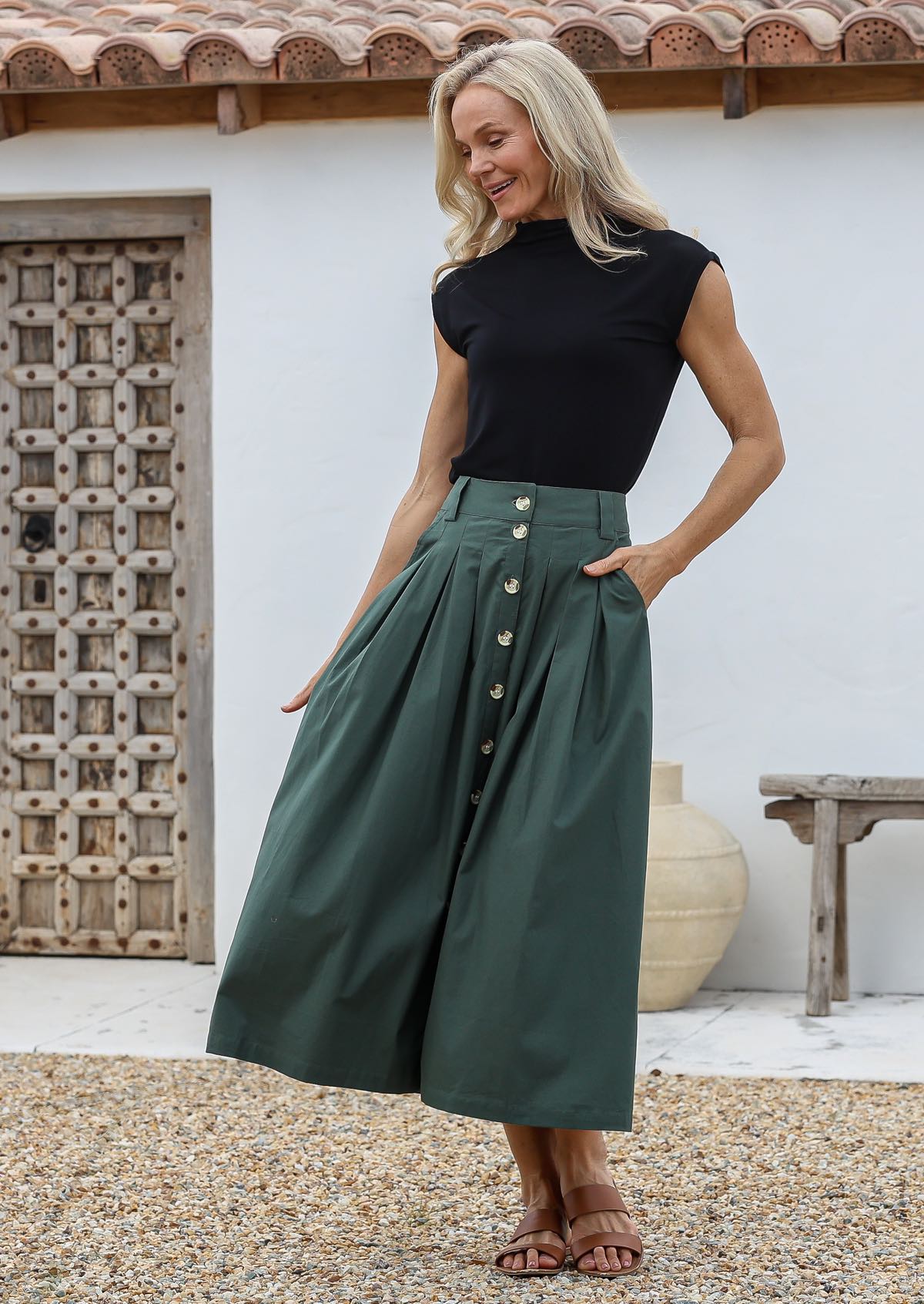 Woman standing with one hand in pocket wearing a 100% cotton shin length pleated skirt in jungle green shade with button up front, box pleats and belt loops