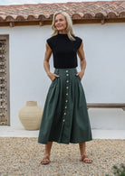 Woman wearing a black top and jungle green long cotton skirt with button through front and box pleats, standing outdoors with a white wall in the background.