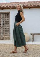 Woman posing sideways with hand in pocket wears 100% cotton pleated skirt in green with belt loops and button through front