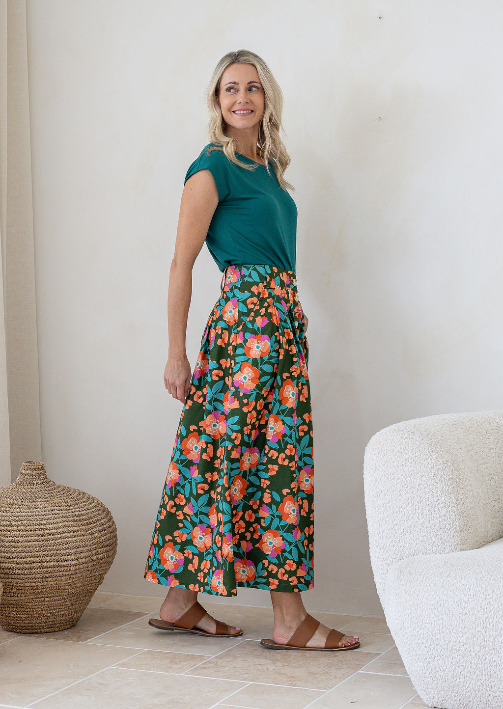 Woman stands wearing a floral print, shin length cotton skirt with button through front and box pleats.