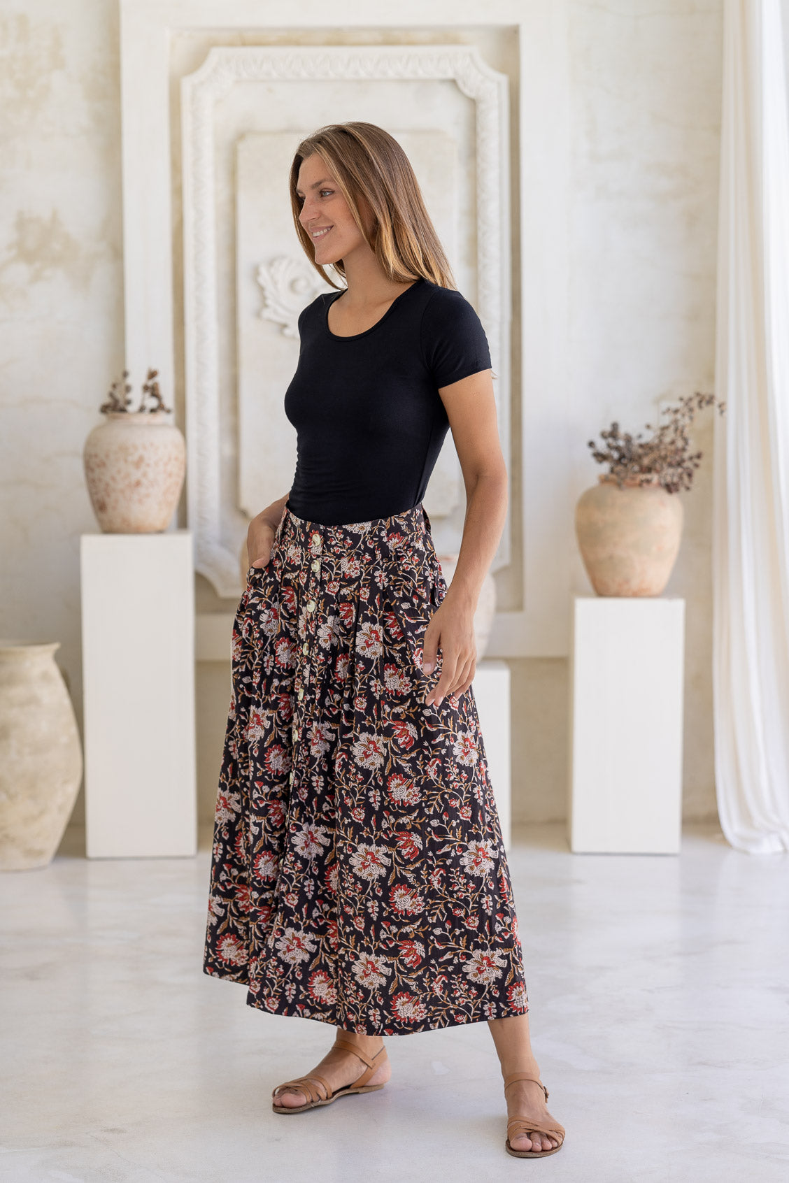 Woman standing with a hand in pocket in a minimalistic room wears 100% cotton button through skirt in black based rust, sand and tan floral prints.