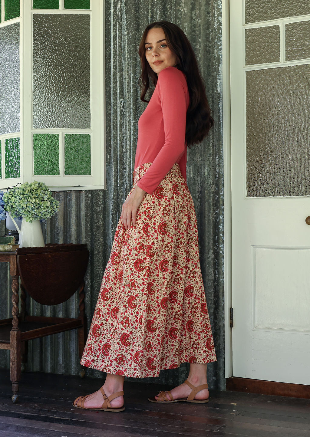 Woman stands sideways wearing a pink top and floral print, shin length cotton skirt with button through front and box pleats.