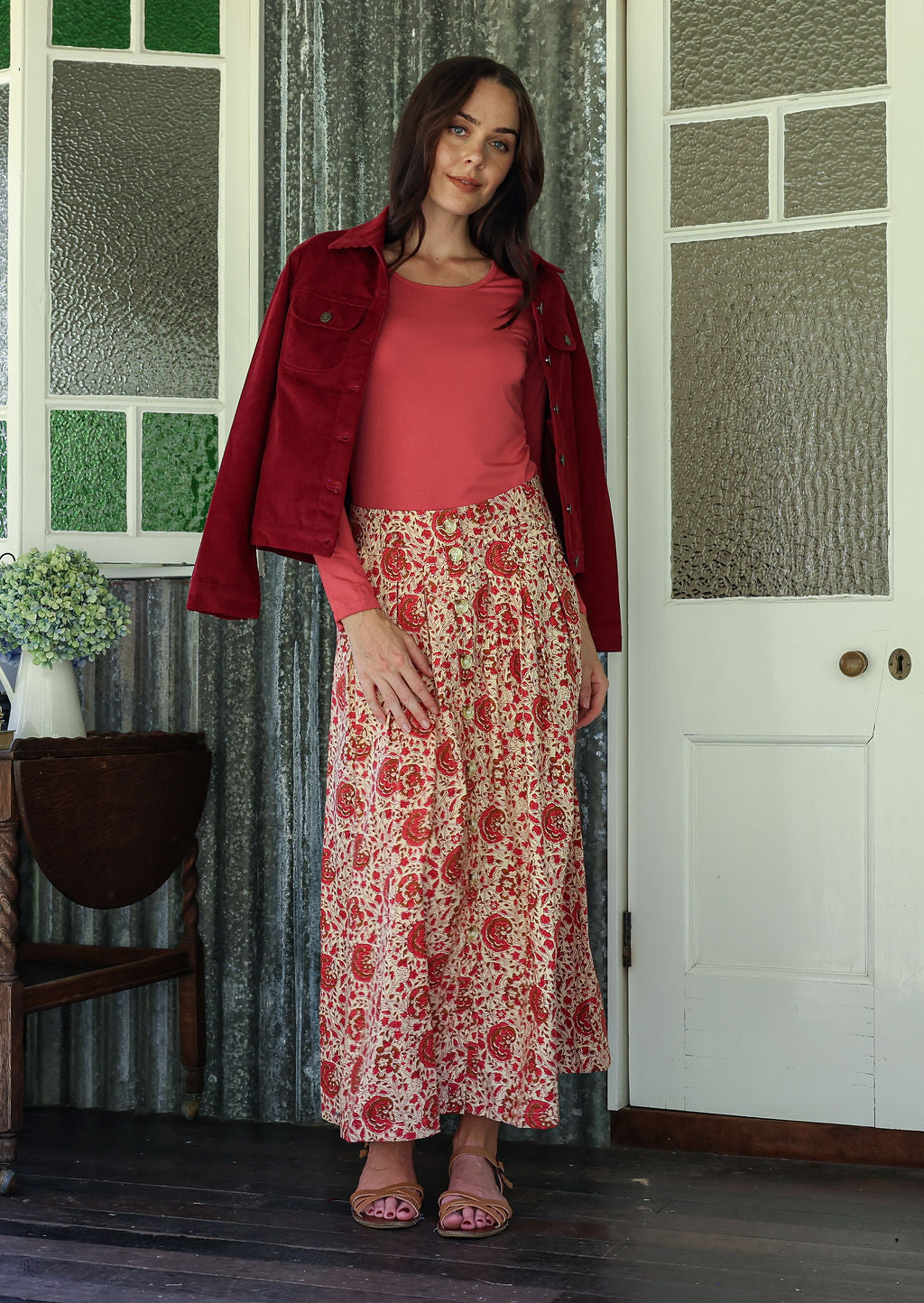 Woman in a red jacket and floral skirt standing in a room with a door and window.