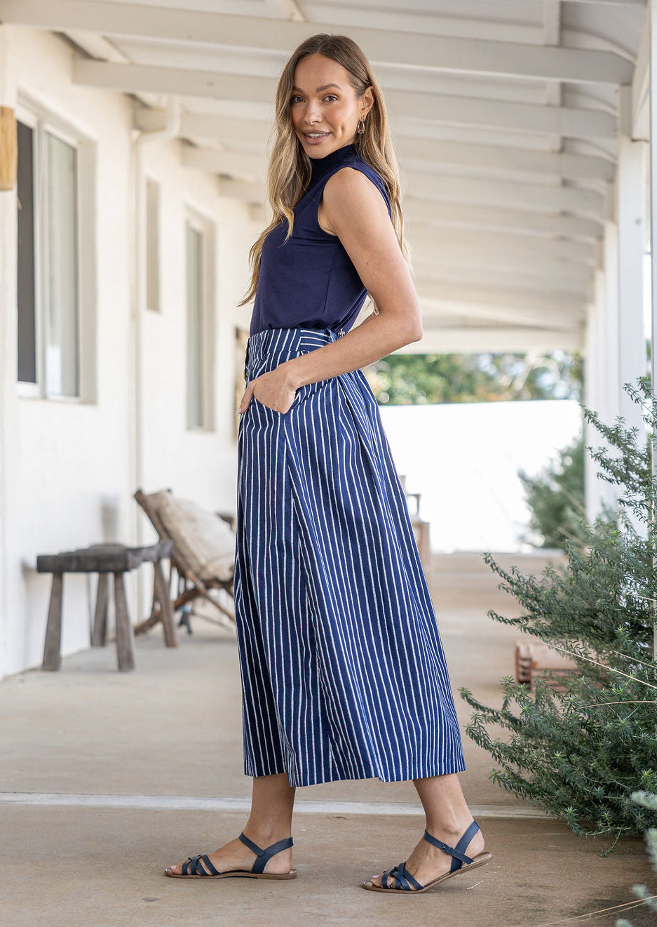 Woman stands sideways wearing a navy top and white striped on navy base print, shin length cotton skirt with button through front and box pleats.