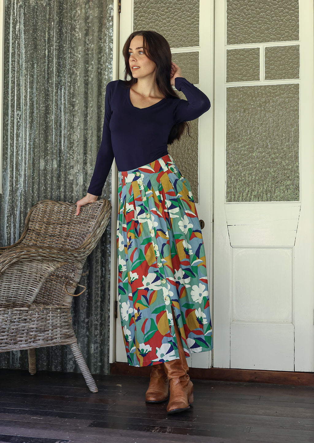 Woman wearing a colorful skirt and navy top standing in a room with wicker chair and patterned walls.