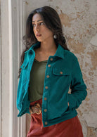 Woman wearing teal corduroy jacket with breast and front pockets, and brass buttons with hands in pockets