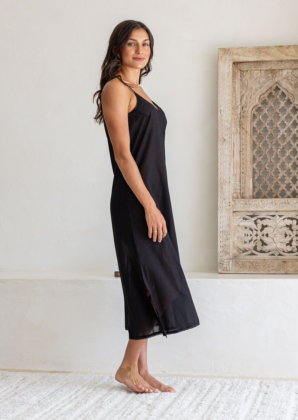 Woman stands side on wearing black cotton voile maxi slip with V-neckline, adjustable spaghetti straps and side splits