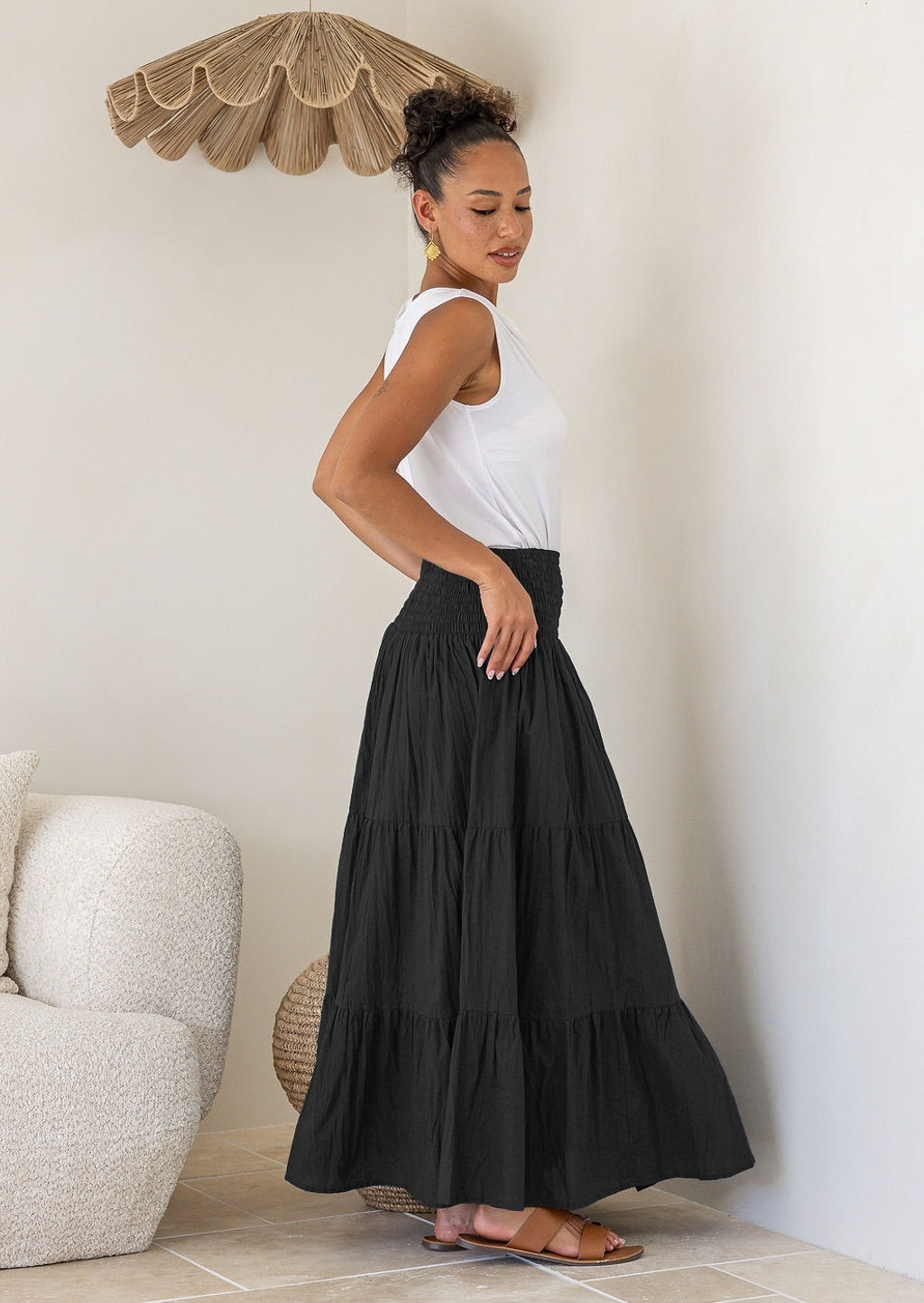 Woman standing side on wearing a white singlet top and black cotton tiered maxi skirt with wide ruched waistband