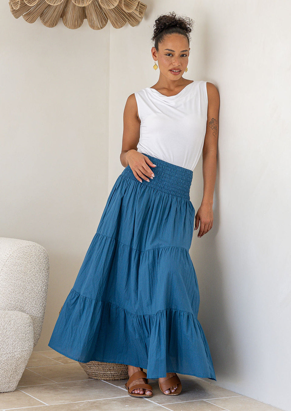 Woman wearing a white singlet top and blue tiered cotton maxi skirt with a wide shirred waistband, leaning against a wall