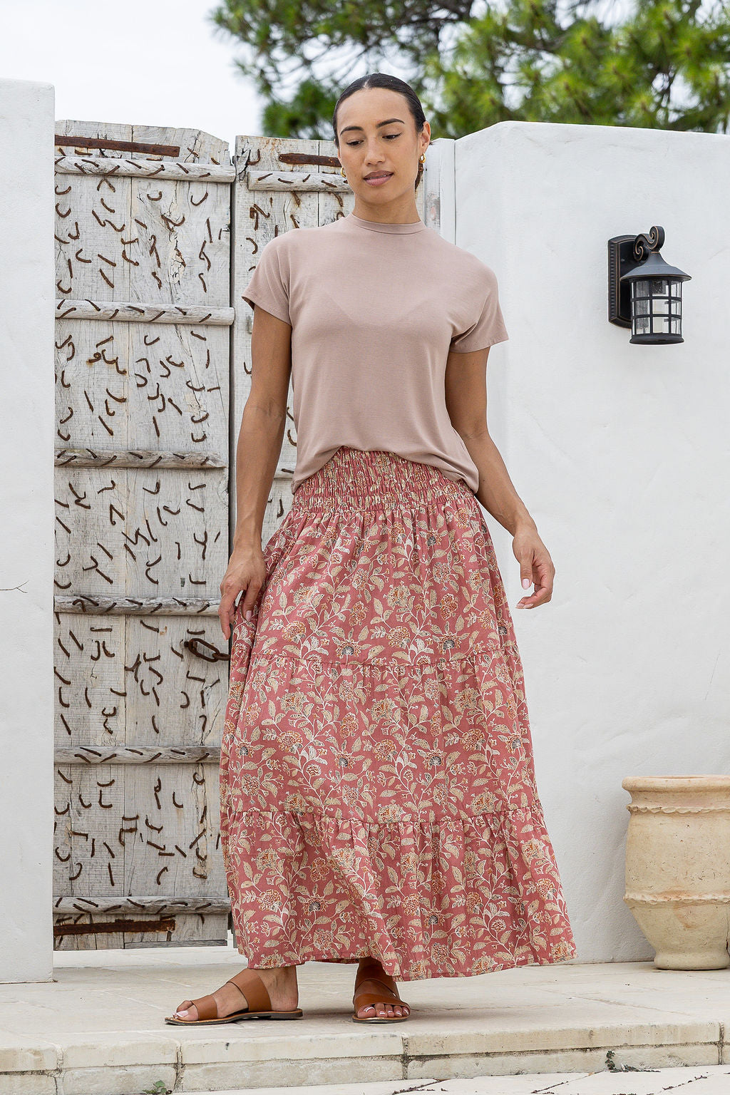Woman wearing a pink top and rose pink based floral skirt standing outdoors against a white wall.