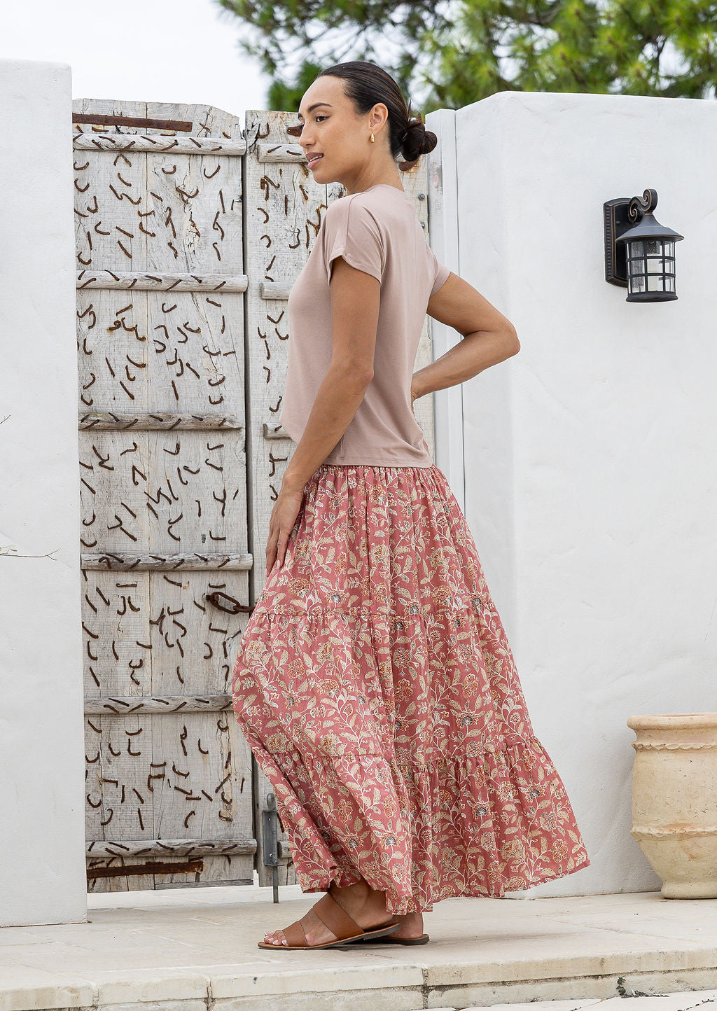 Woman wearing a pink top and floral skirt standing in front of a white wall with a decorative pattern.