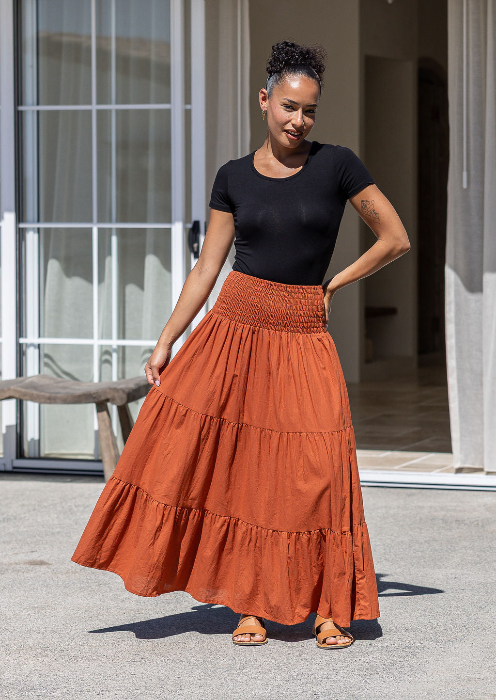 Woman wearing a black top and rust-coloured tiered cotton maxi skirt standing holding the skirt out to the side