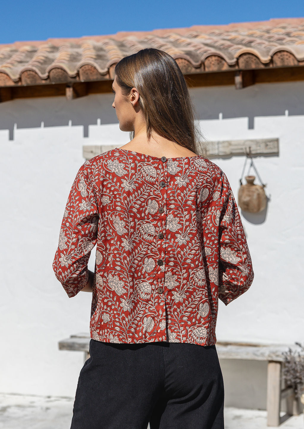 Woman stands showing the back of the cotton top with decorative buttons down the centre of the back.