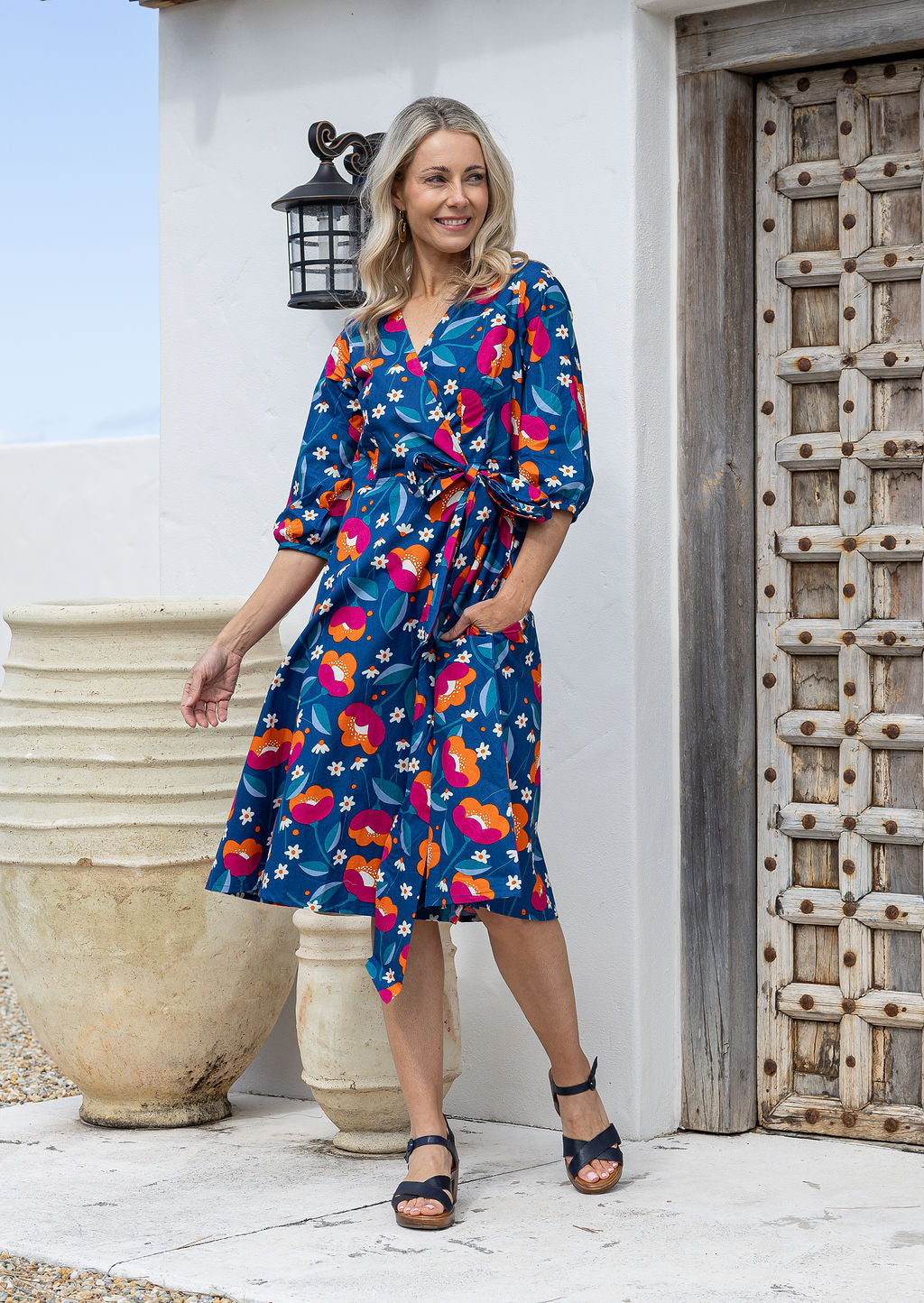 Model wears a royal blue based floral printed cotton, over the knee length wrap dress with V-neckline, 3/4 sleeves, and a wide waist-tie