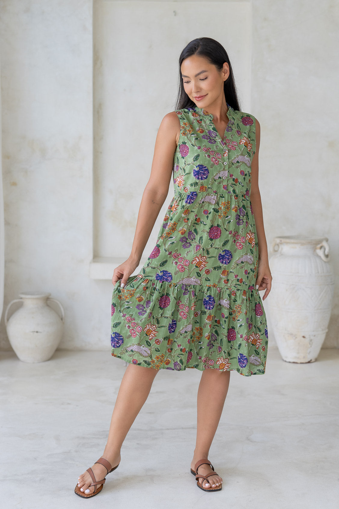 Woman holding skirt out to the side of a green floral sleeveless, knee length dress with a buttoned bodice, tiered skirt and pockets.