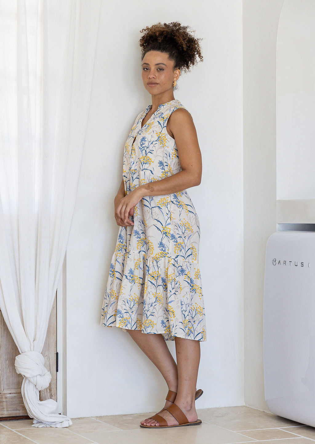 Woman stands sideways wearing a sleeveless neutral floral cotton dress with pockets, mandarin collar and a two tiered skirt.