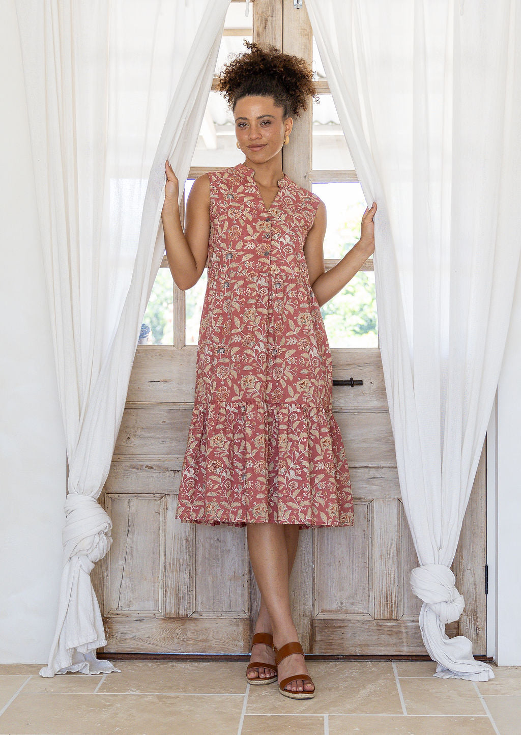 Woman wearing a rose pink based floral sleeveless, knee length dress with a buttoned bodice, tiered skirt and pockets.