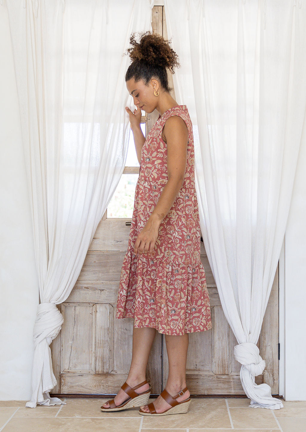 Woman stands sideways wearing a sleeveless rose pink floral cotton dress with pockets, mandarin collar and a two tiered skirt.