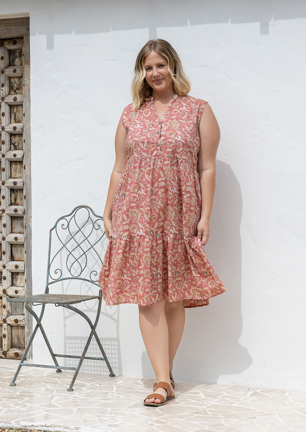 Woman posing in a rose pink based floral sleeveless dress with a buttoned bodice, two tiered skirt and mandarin collar