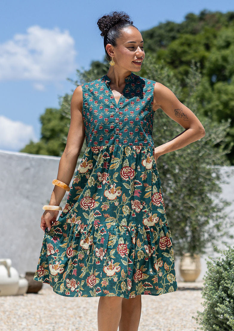 Woman wearing a knee length cotton sleeveless dress with buttoned bodice and two tiered skirt in green based floral print.