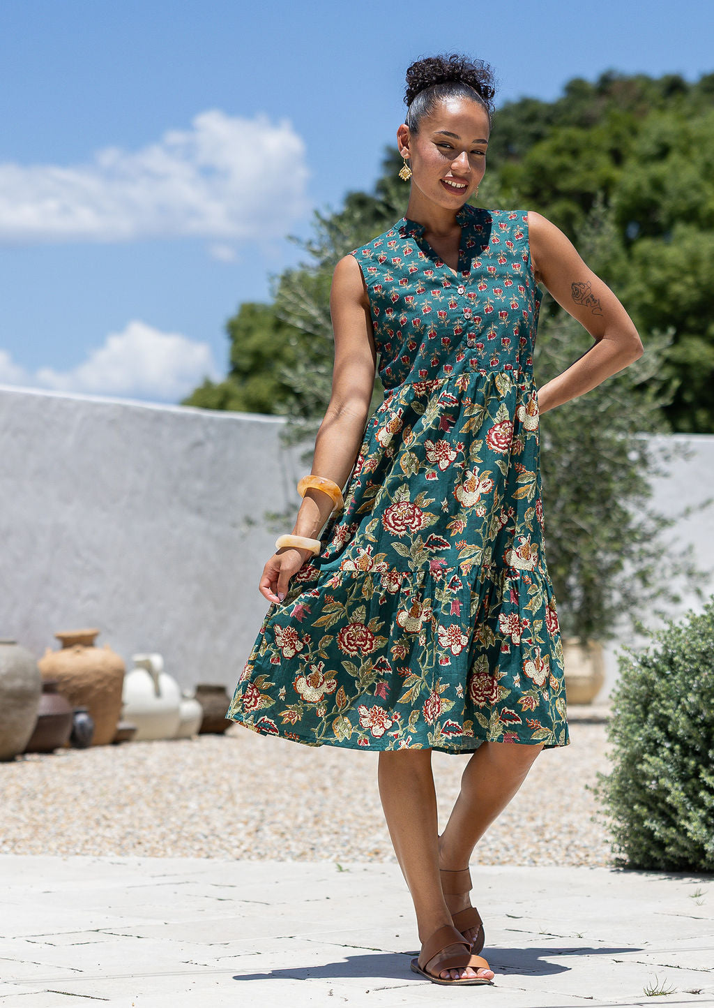 Woman holding skirt out to the side of a green floral sleeveless, knee length dress with a buttoned bodice, tiered skirt and pockets.