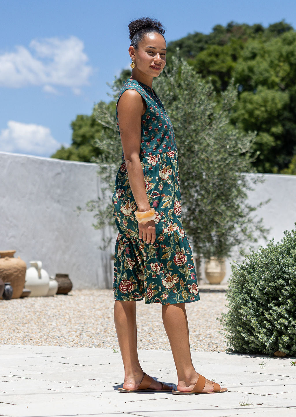 Woman stands sideways wearing a sleeveless green floral cotton dress with pockets, mandarin collar and a two tiered skirt.