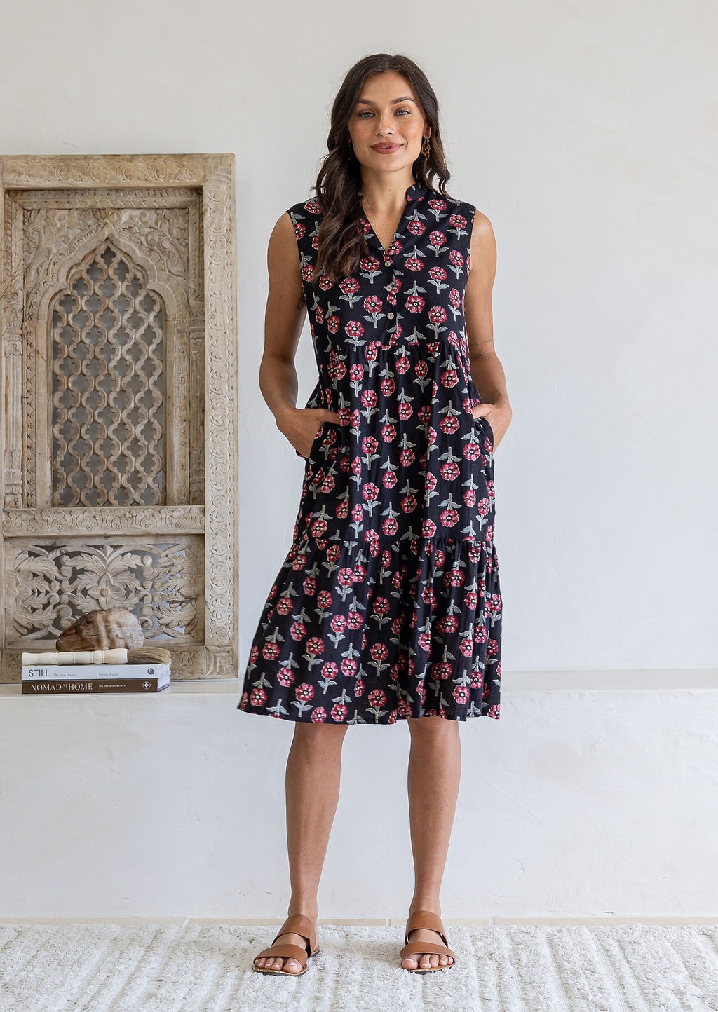 Woman wearing a knee length cotton sleeveless dress with buttoned bodice and two tiered skirt inn earthy black based floral print.