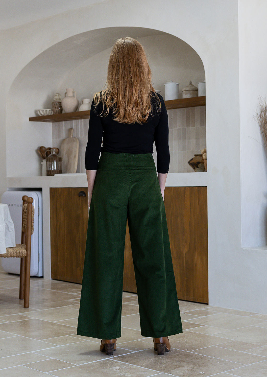 Woman stands backwards wearing a cotton corduroy wide leg pants in olive green paired with a basic black top.