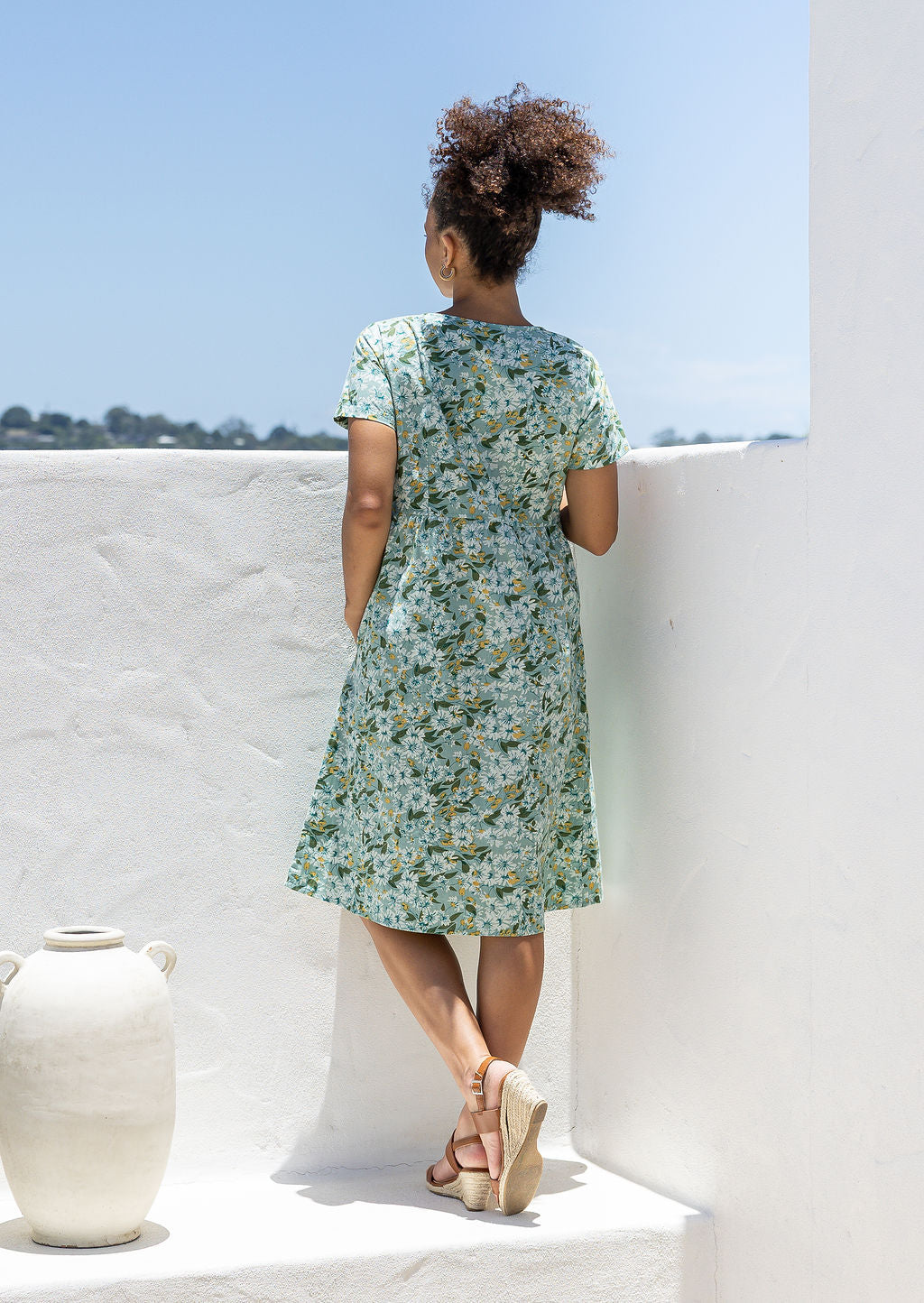 Model stands backwards wearing a cotton V-neck relaxed fit dress with empire waistline and short sleeves.