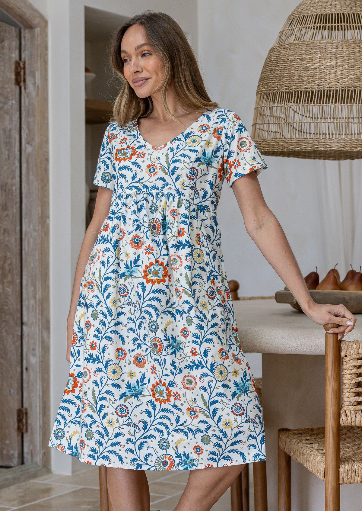 Woman poses in cotton over the knee length relaxed fit dress with short sleeves, V-neckline and empire waistline, in white based floral print in a white dining room