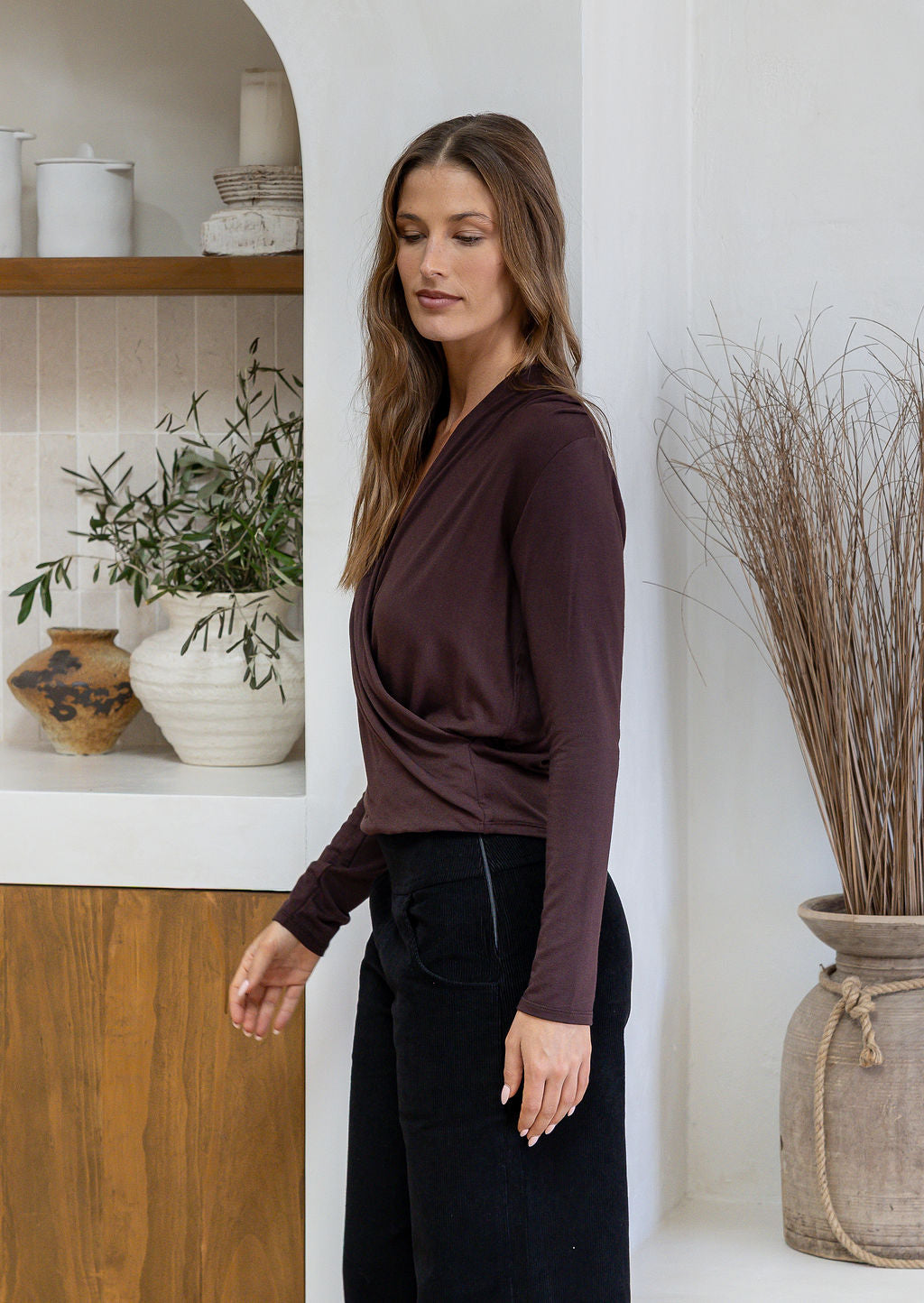 Woman stands side on wearing a false wrap front, long sleeve top in dark brown stretch rayon jersey, paired with black pants